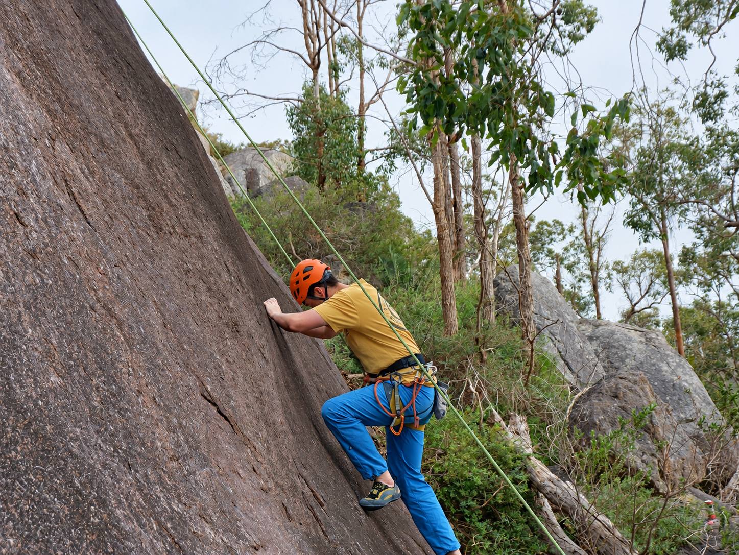 A climber in an orange helmet starting up a granite slab on a top rope, with gum trees and boulders behind.