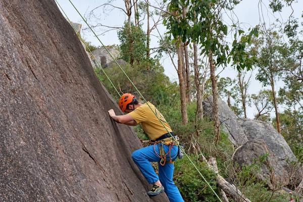 A climber in an orange helmet starting up a granite slab on a top rope, with gum trees and boulders behind.