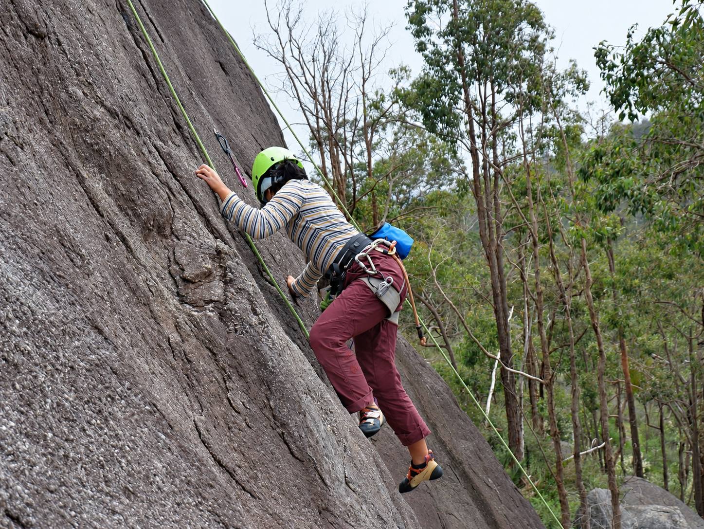 A climber in a green helmet moving up a steep granite slab on a top rope, with gum trees behind.