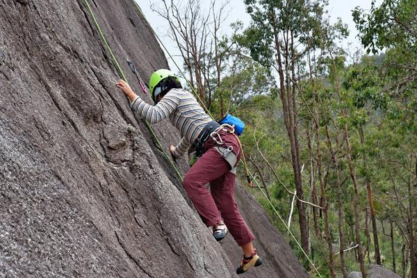 A climber in a green helmet moving up a steep granite slab on a top rope, with gum trees behind.