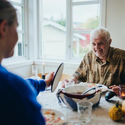 En eldre mann med grått hår og rynker smiler mens han sitter ved et spisebord. Foran ham står en stor gryte med varm mat.