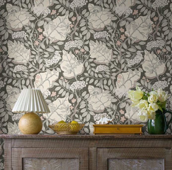 An interior with dark botanical wallpaper featuring cream and pink accents, a wooden cabinet, a lamp, and a vase of white flowers.