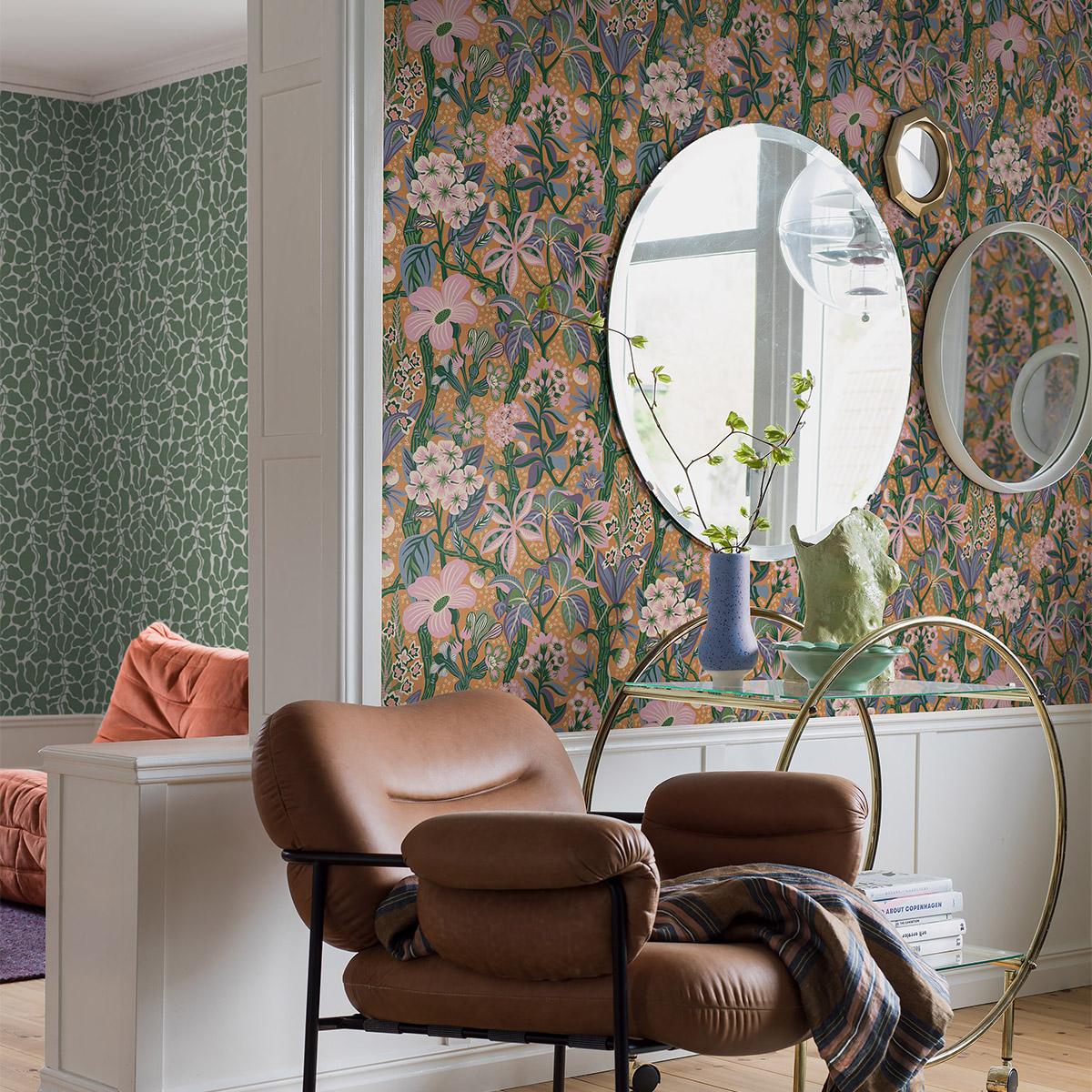 A cozy room with a brown leather armchair, a gold and glass bar cart, and mirrors against a vibrant floral wallpaper, with a green patterned wallpaper visible in an adjacent room.