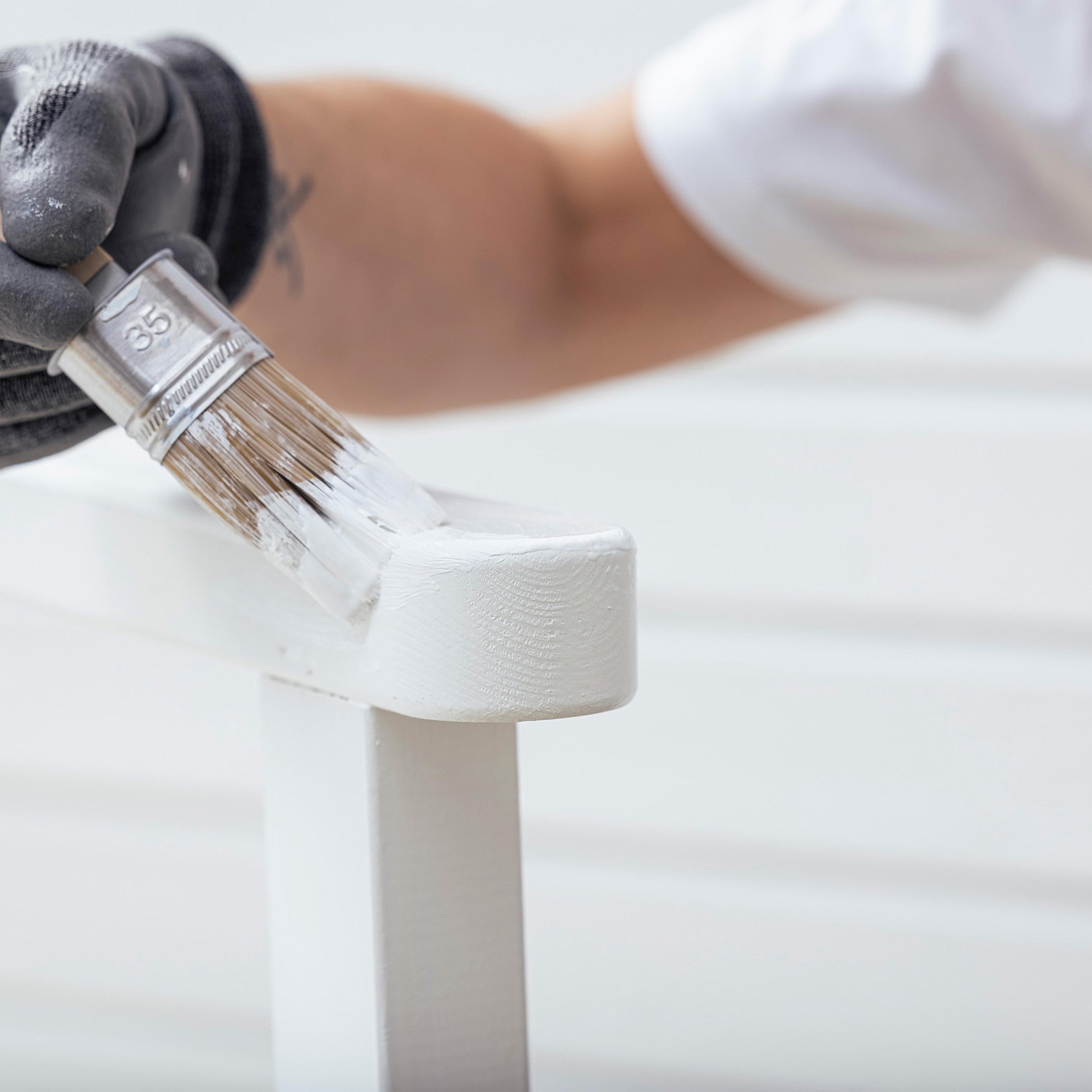 A gloved hand uses a paintbrush to apply white paint to a white object.