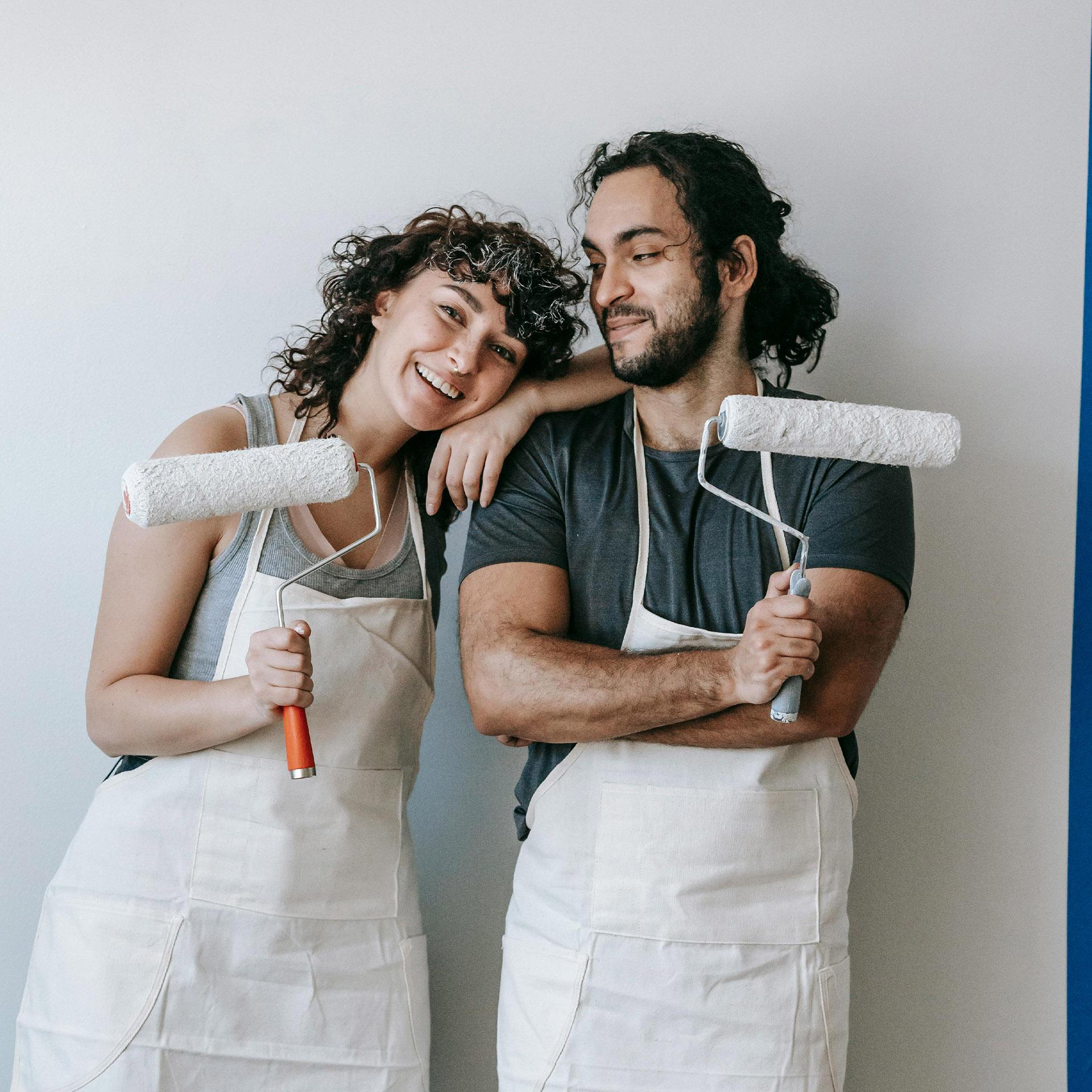 A smiling couple wearing aprons and holding paint rollers.