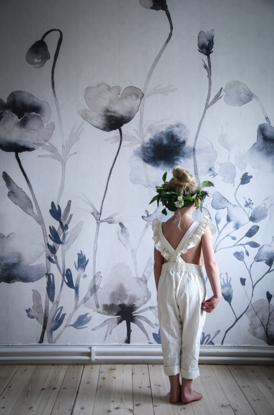 A child wearing a floral crown stands with their back to the viewer, facing a wall with large grey and blue watercolor flower patterns.