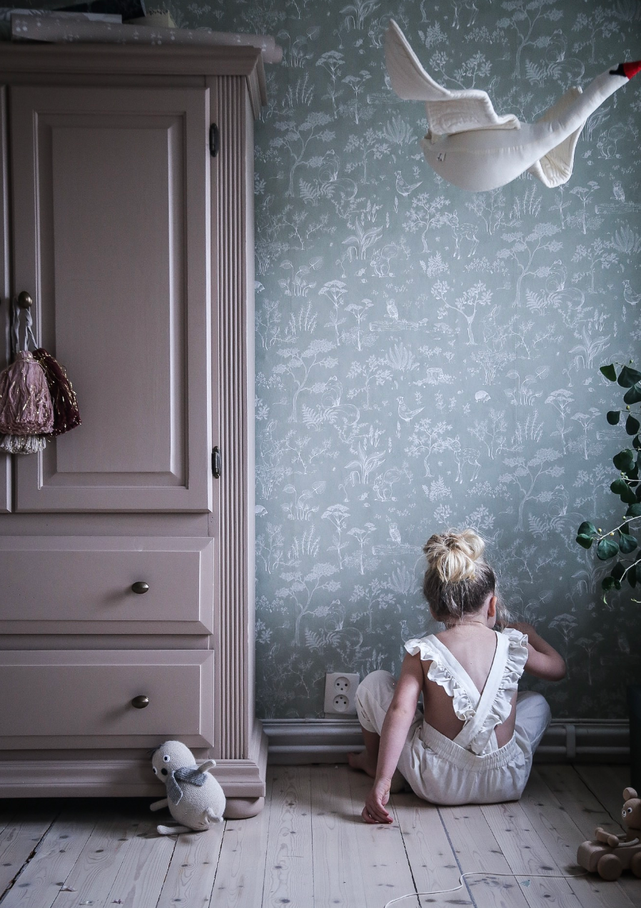 A child sits on a wooden floor with their back to the camera, facing a patterned wall next to a large armoire and a mounted swan decoration.