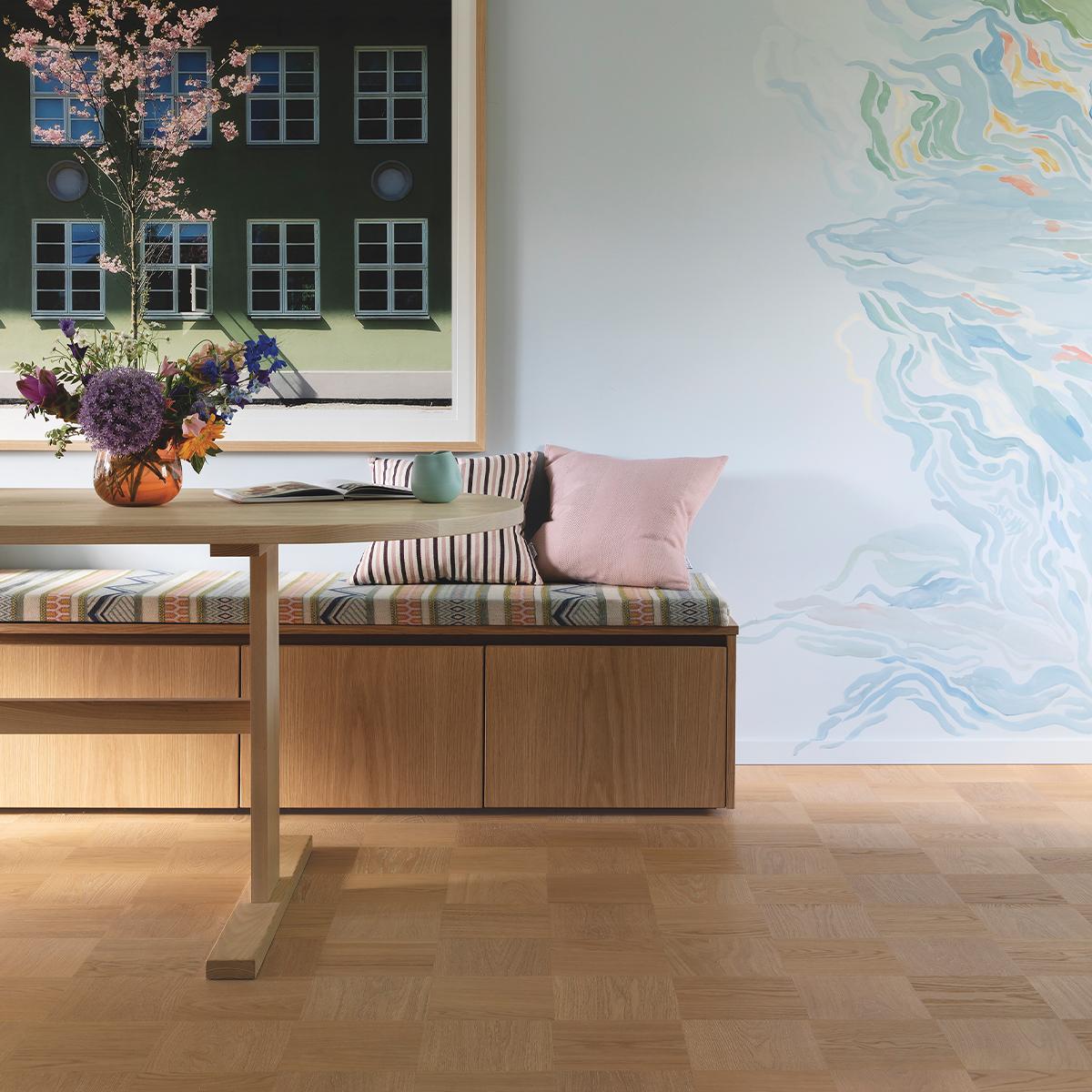 A modern room with a light wooden table and a built-in bench with patterned cushions. A framed building print and a blue watercolor mural adorn the walls, above a parquet floor.