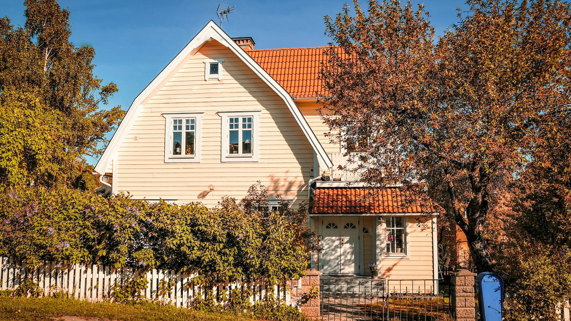 Light yellow house with a red roof, surrounded by autumn trees and a white picket fence under a clear blue sky.