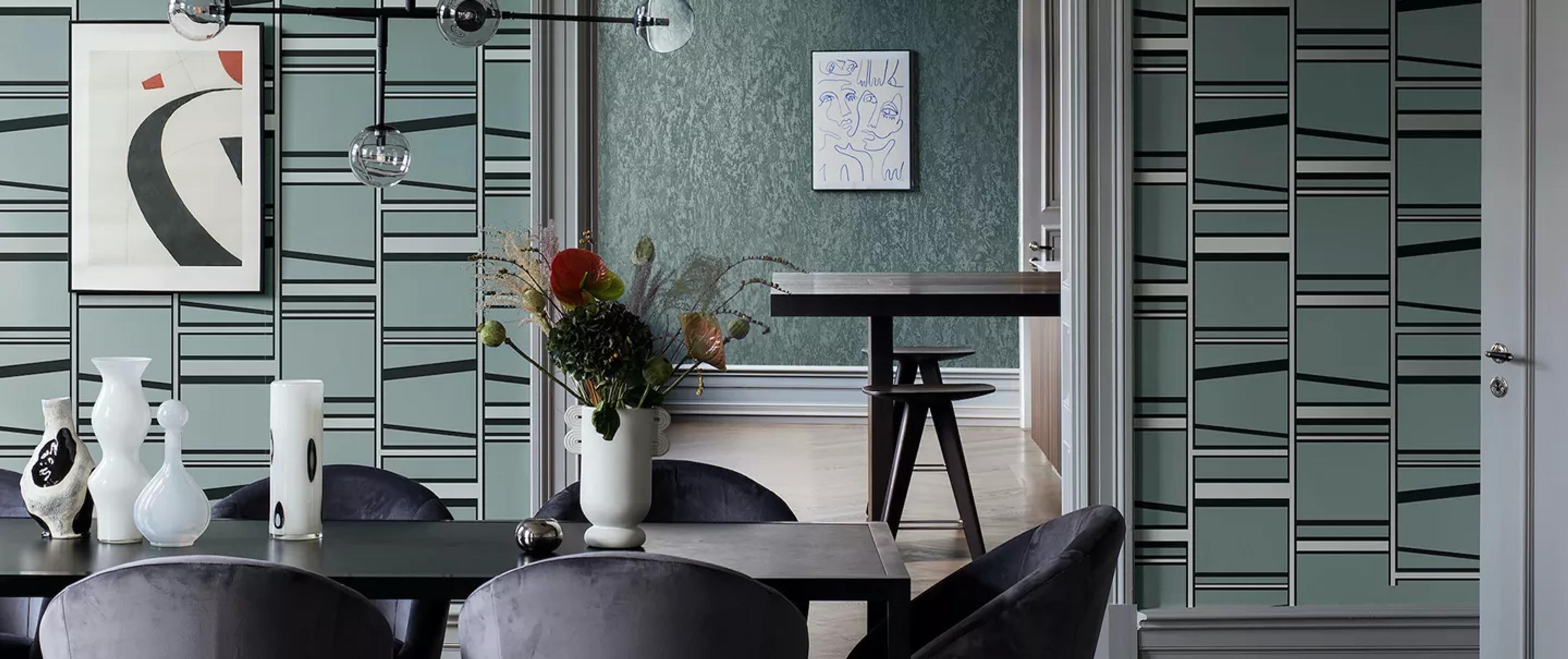 A modern dining room with teal geometric wallpaper, a long dark dining table, dark velvet chairs, and a flower arrangement.