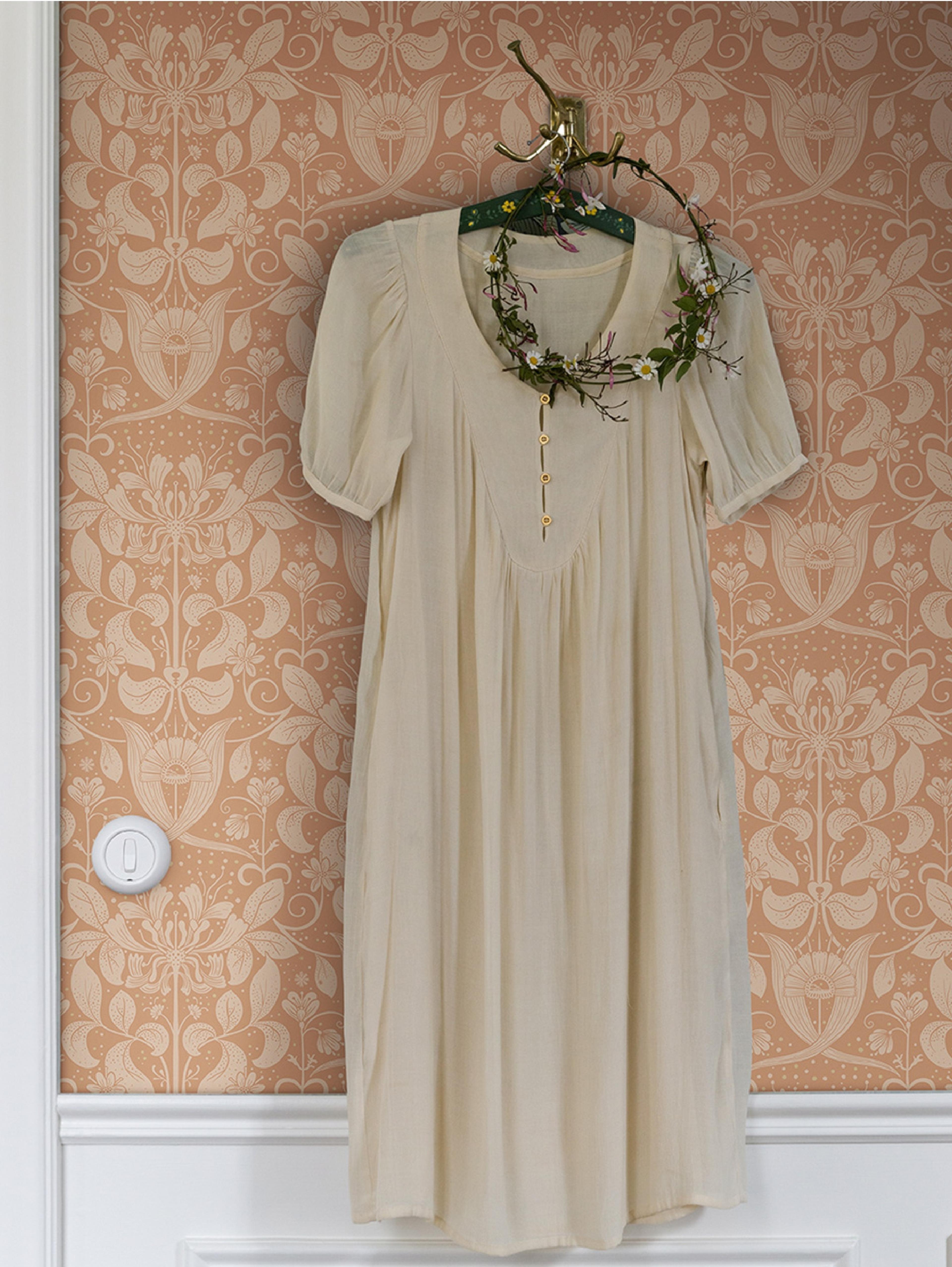 A cream dress with a floral wreath hangs on a hook against peach patterned wallpaper.