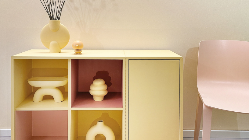 Pale yellow modular cabinet with pink accents displaying decorative objects, next to a pale pink chair.