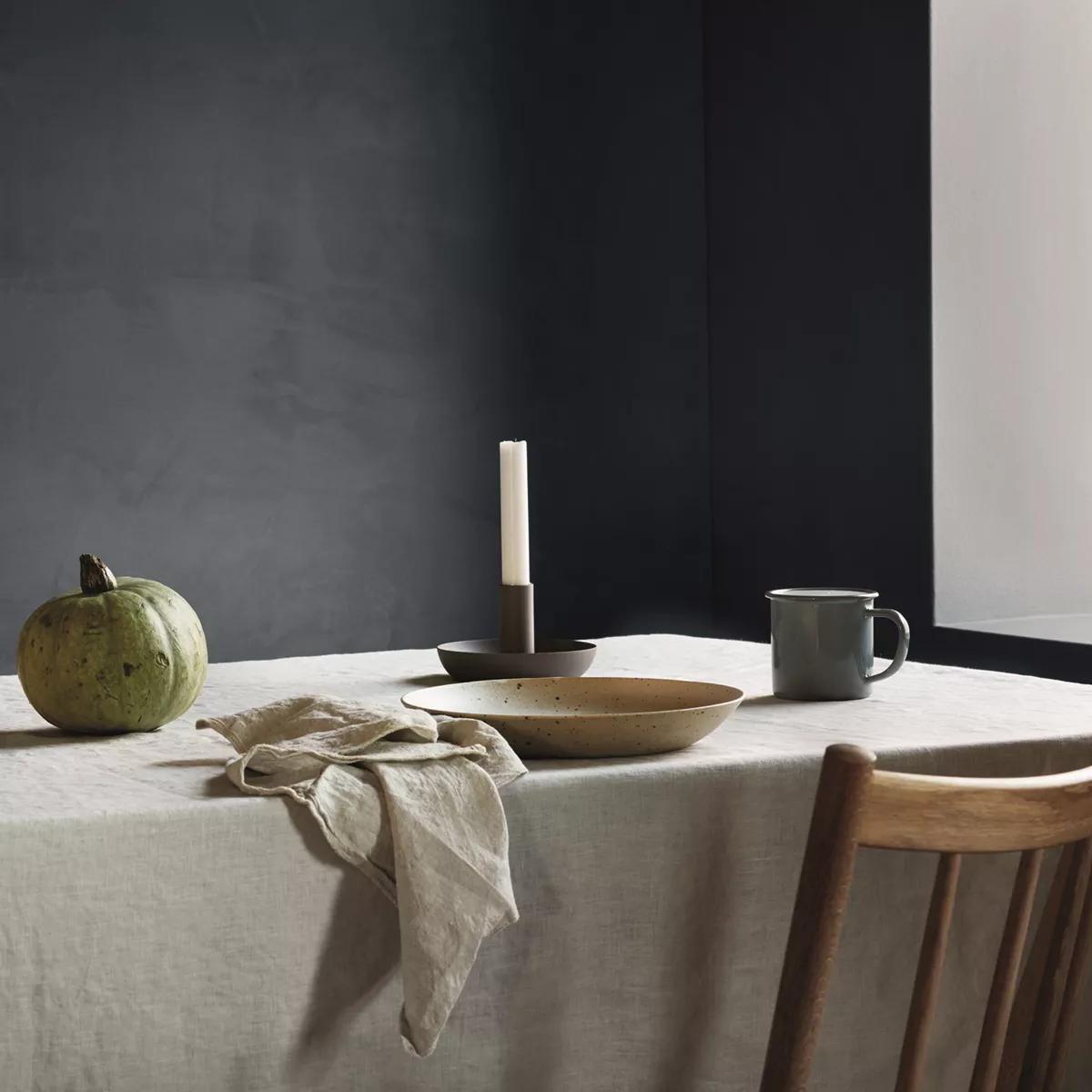 A table with a green pumpkin, candle, ceramic bowl, linen napkin, and mug on a light linen tablecloth.
