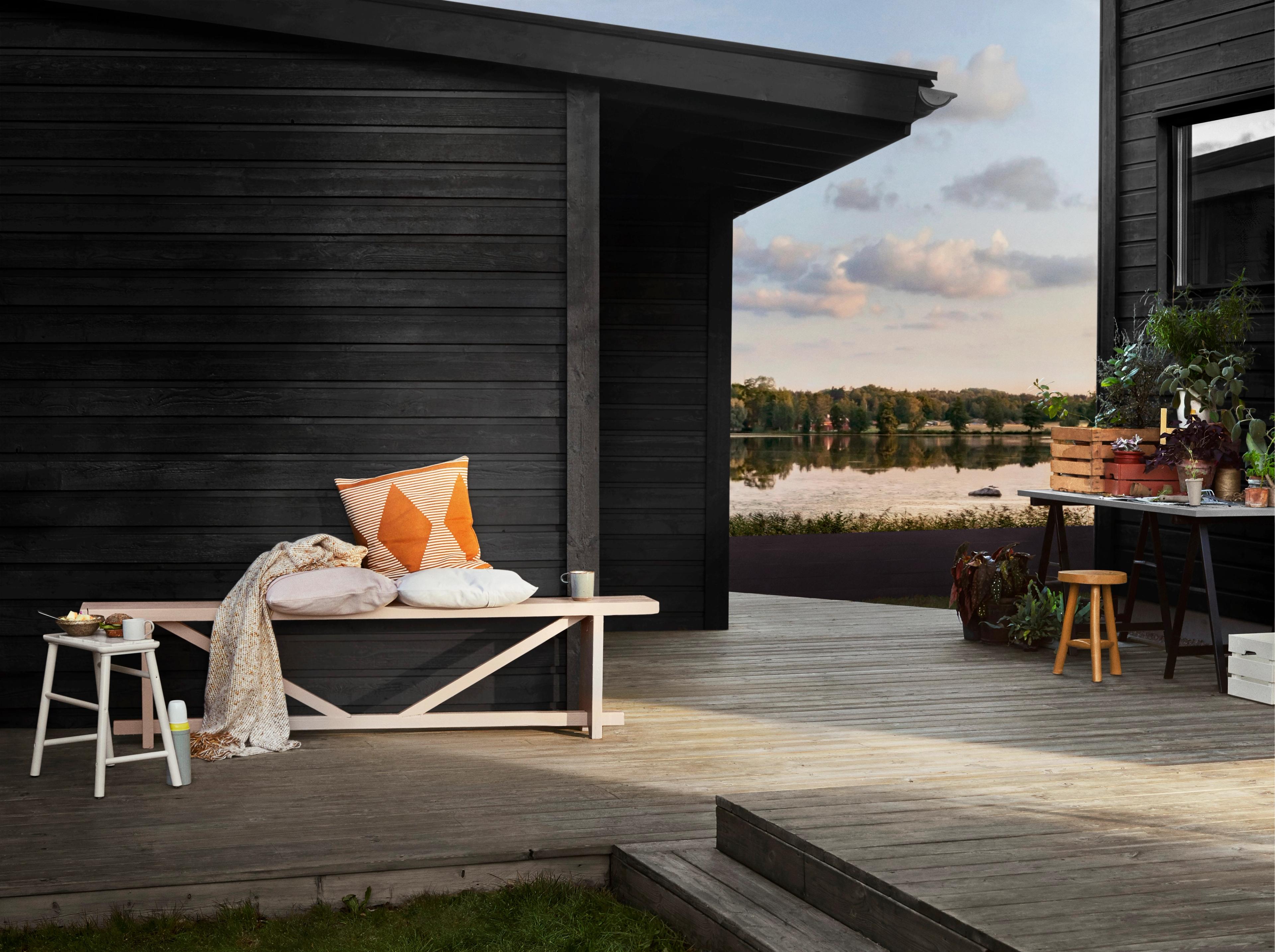 Black wooden cabin with a light pink bench on a wooden deck overlooking a lake, with potted plants on a table.
