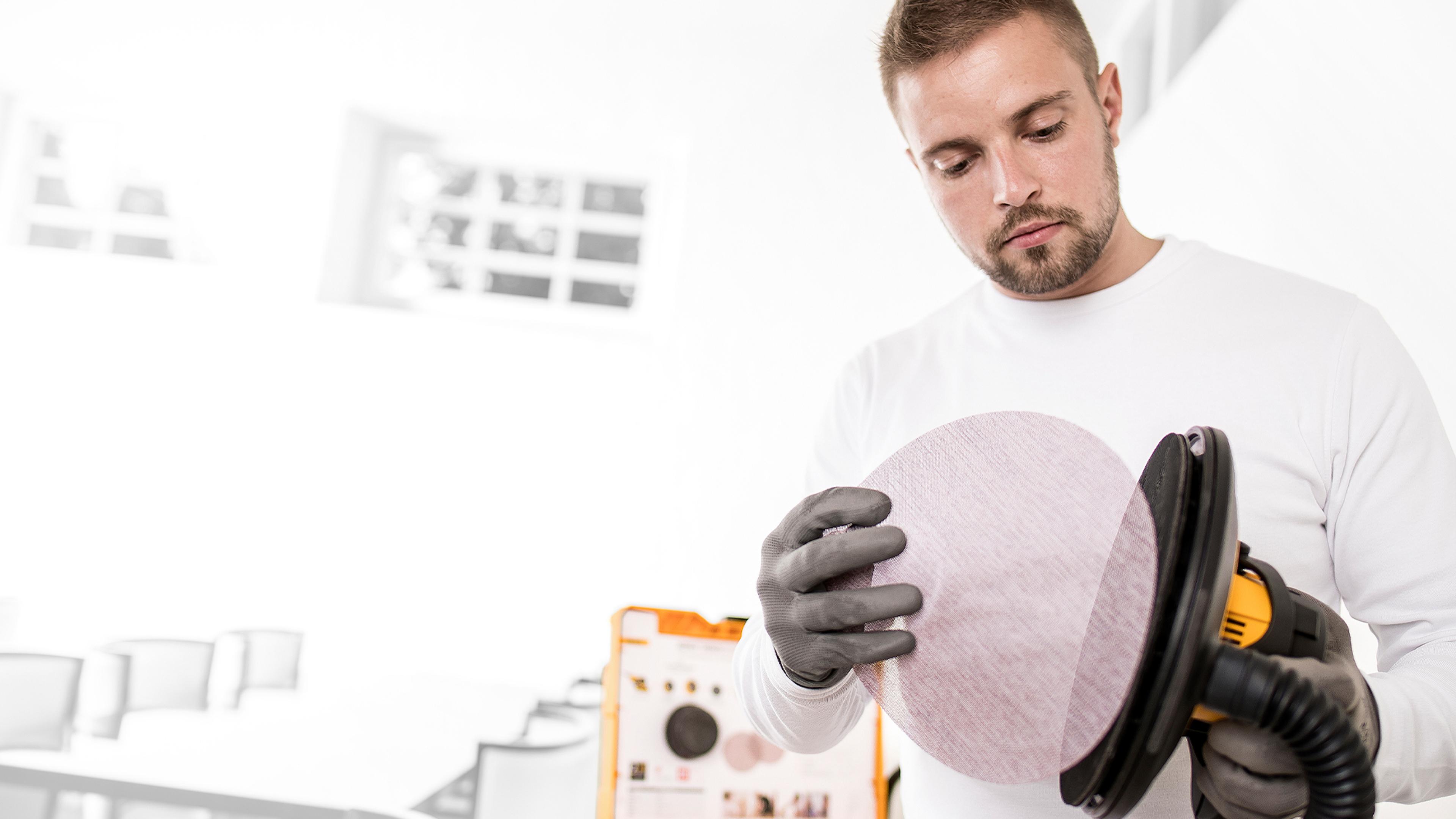 A man in a white shirt and gloves attaches a round sanding disc to a power sander.