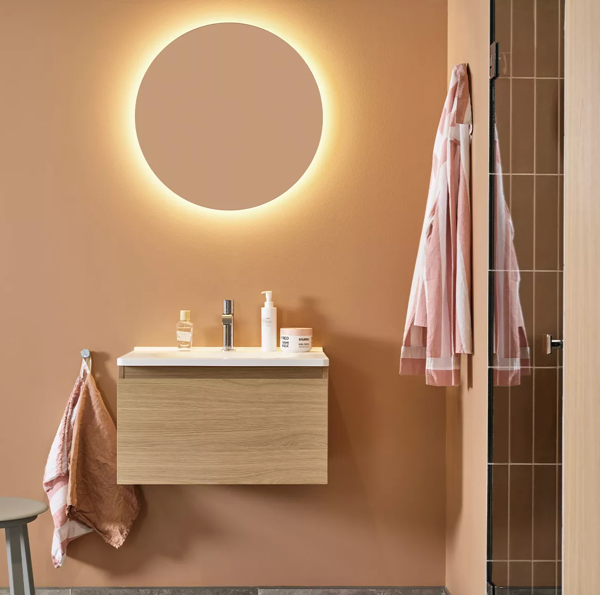 A modern bathroom with a round illuminated mirror above a wooden vanity and white sink on a warm brown wall, with towels and a shower.