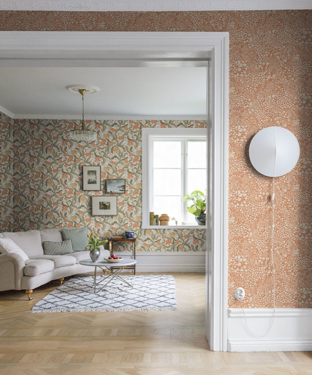 A view through a doorway connecting two rooms, one with reddish-orange botanical wallpaper and a wall lamp, the other with green botanical wallpaper and a sofa.