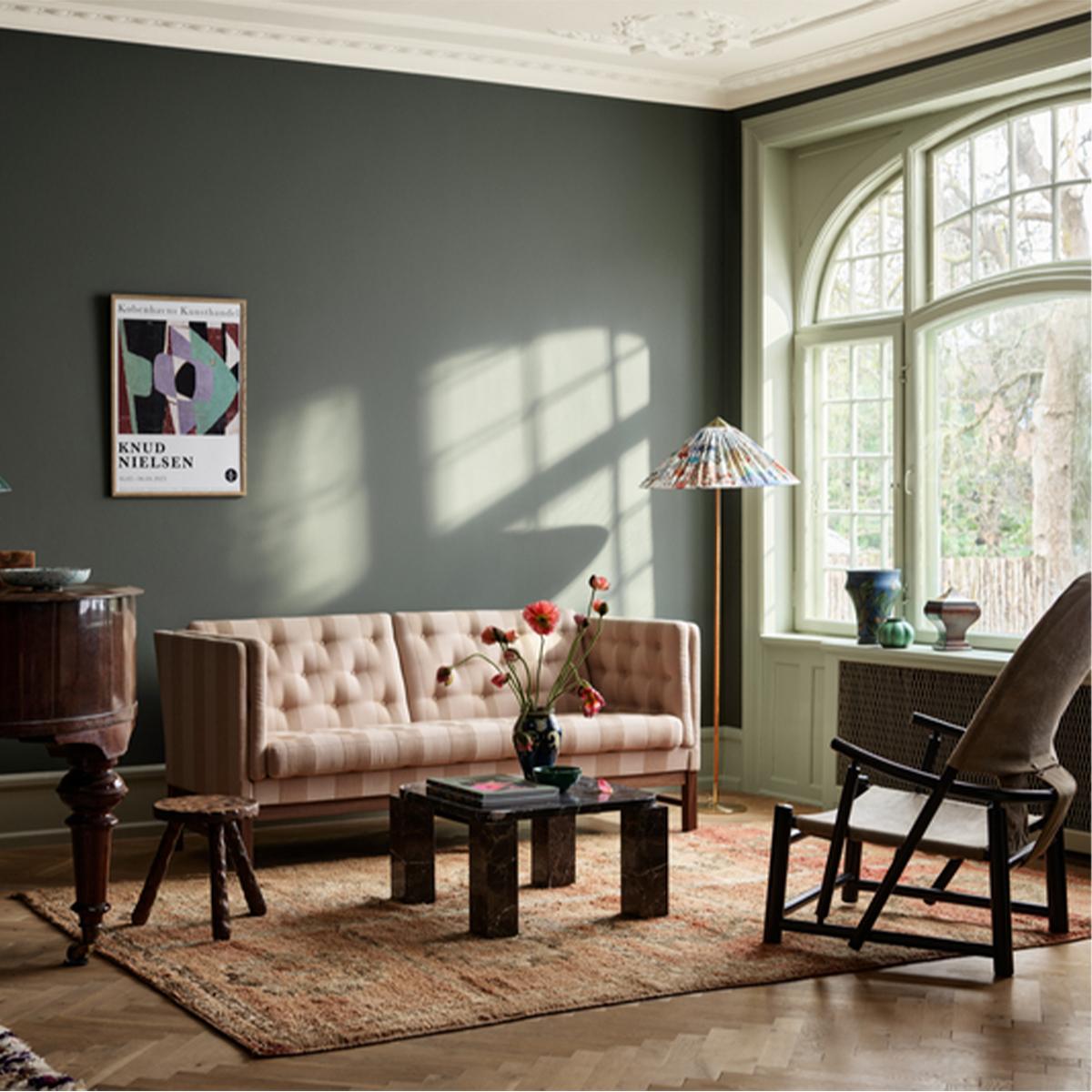 A dark green living room with a pink tufted sofa, marble coffee table, colorful pleated floor lamp, modern armchair, and a large arched window.