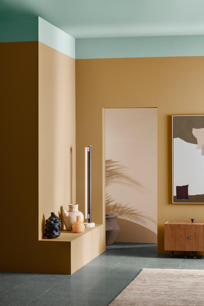 A stylish room with warm ochre walls, a mint green ceiling, and dark grey floors. A modern lamp and decorative vases sit on a built-in ledge, while an abstract painting and wooden cabinet adorn the right wall.