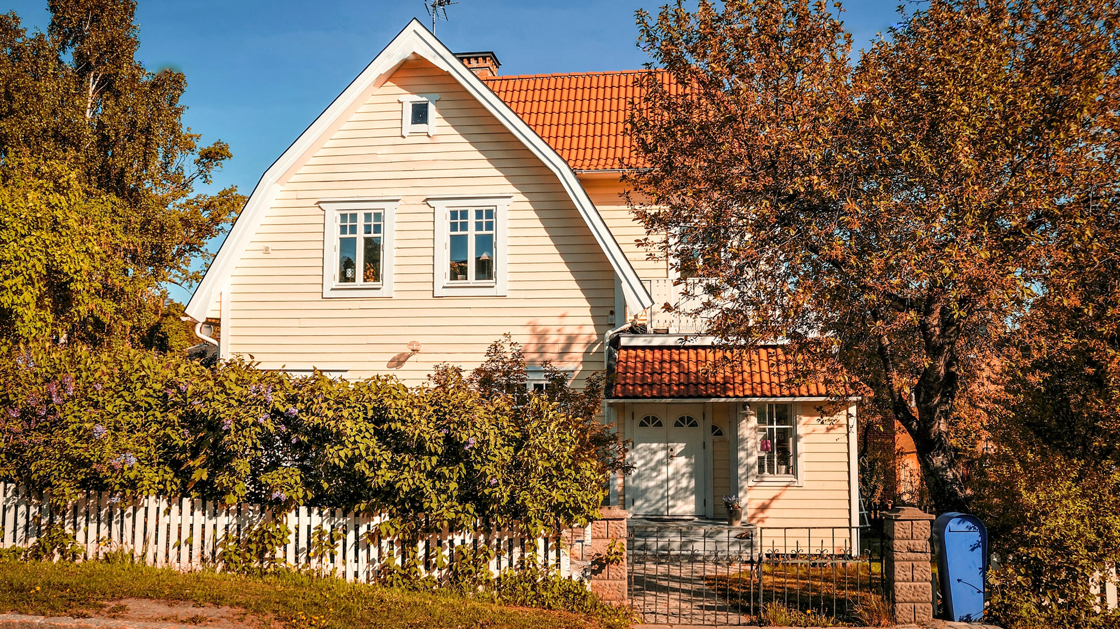 A light yellow house with an orange tiled roof behind a white picket fence, surrounded by lush bushes and autumn trees under a bright blue sky.