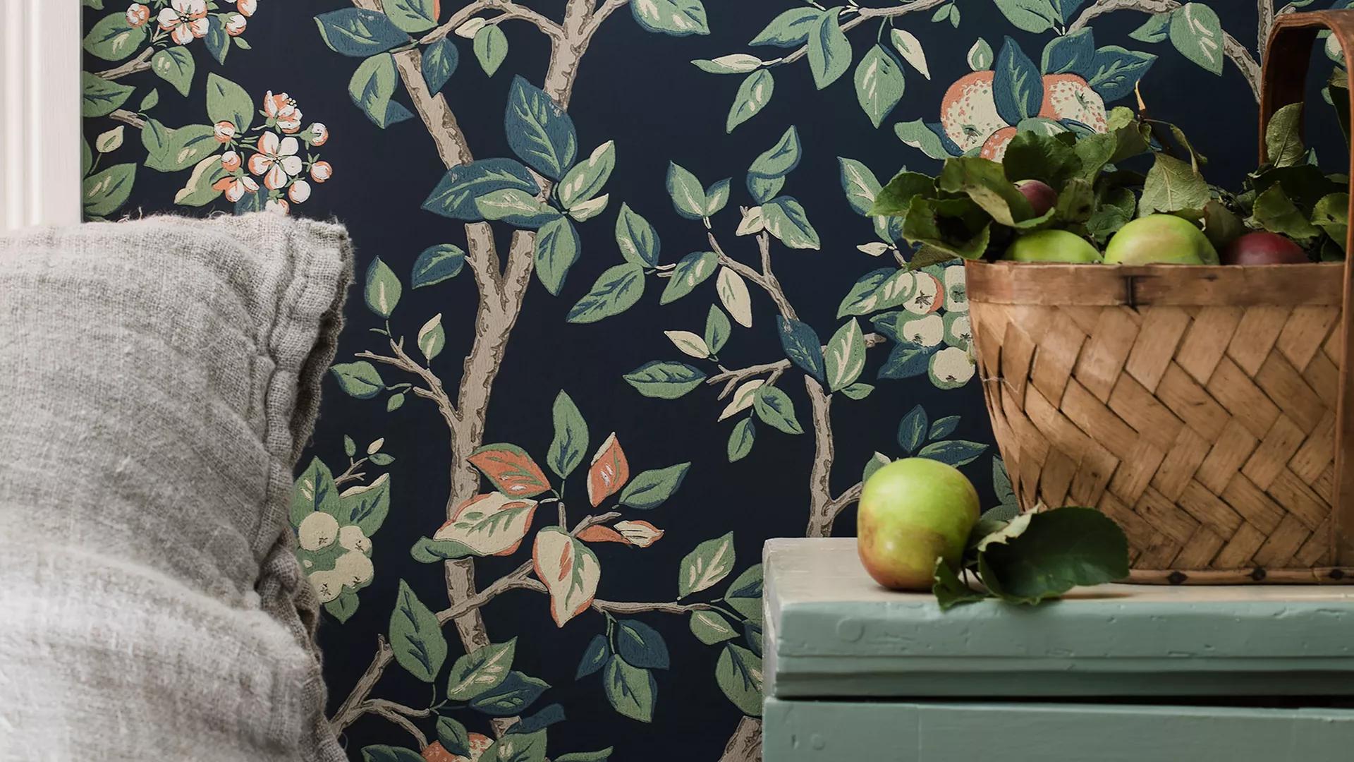 Dark blue wallpaper with an apple tree pattern, a grey pillow, and a basket of apples on a teal surface.