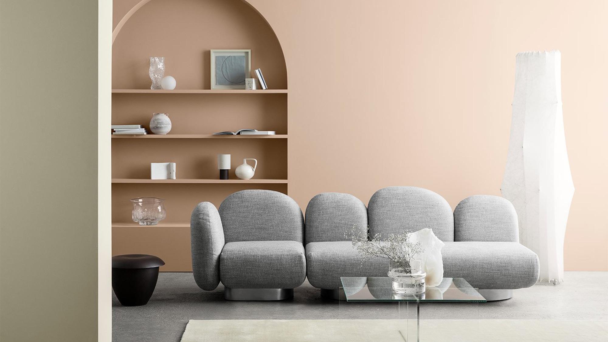 A modern, minimalist living room with a grey modular sofa, glass coffee table, and an arched built-in bookshelf displaying decor, set against light peach and sage green walls.