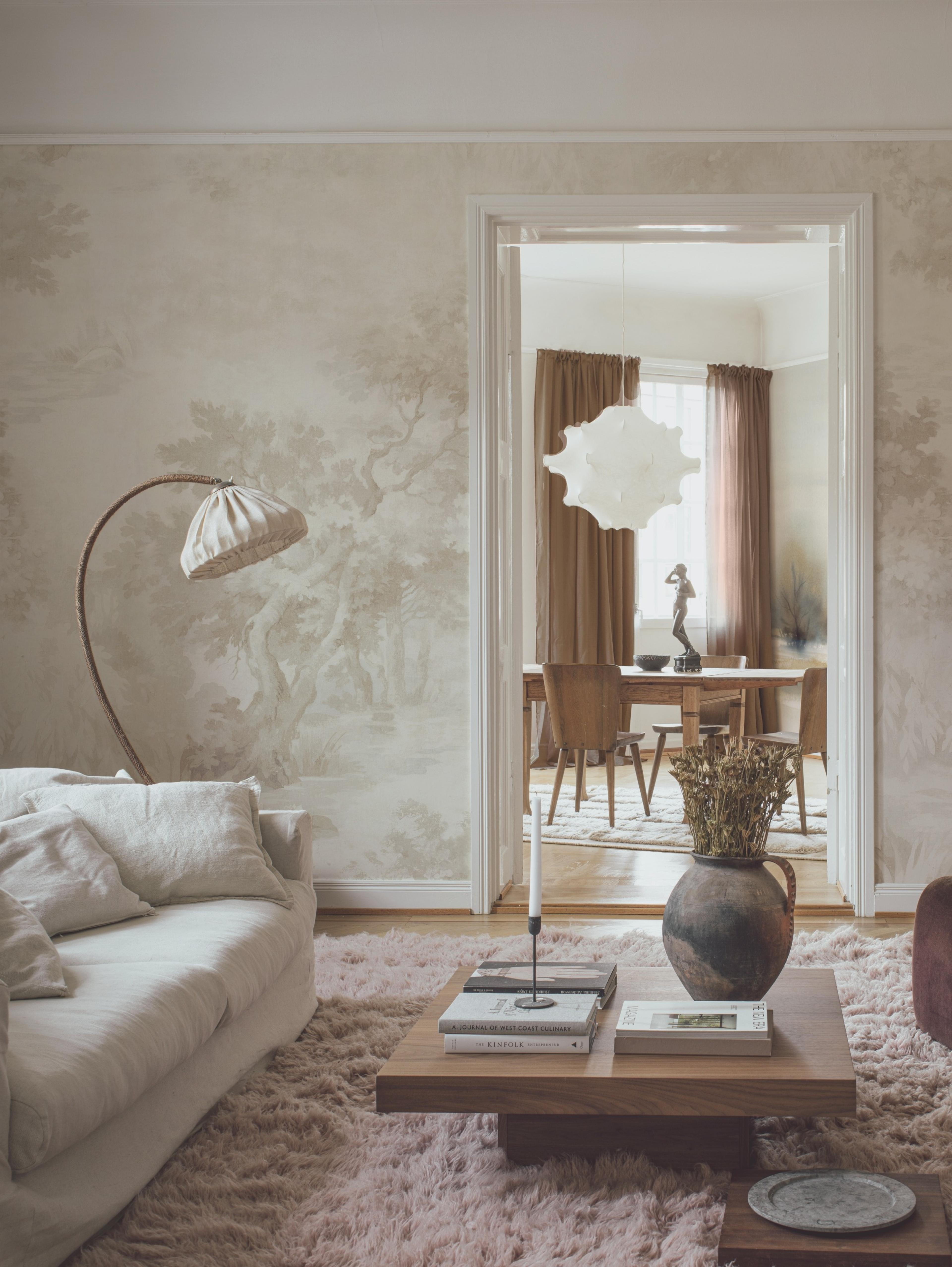 A living room with light beige landscape wallpaper, a white sofa, a pink shag rug, and a wooden coffee table displaying books and dried flowers, with an open doorway to a dining room.
