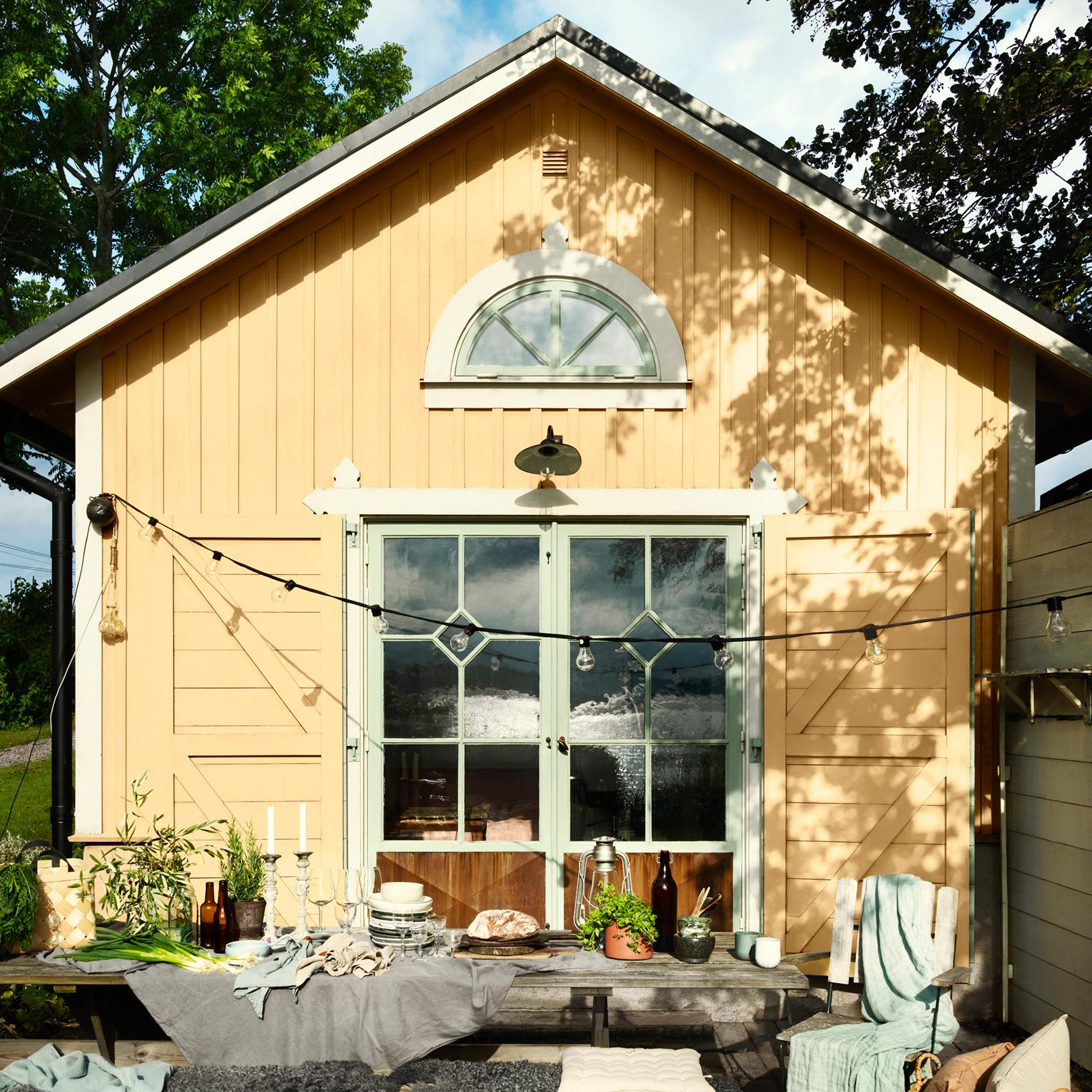A rustic yellow wooden building with green-framed windows and doors, featuring an outdoor dining table set with food and string lights.