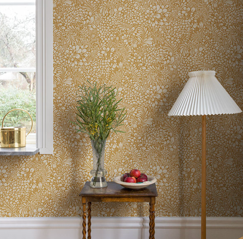A room with mustard yellow and white floral wallpaper, a window, a brass watering can, a wooden table with green foliage and apples, and a pleated white floor lamp.
