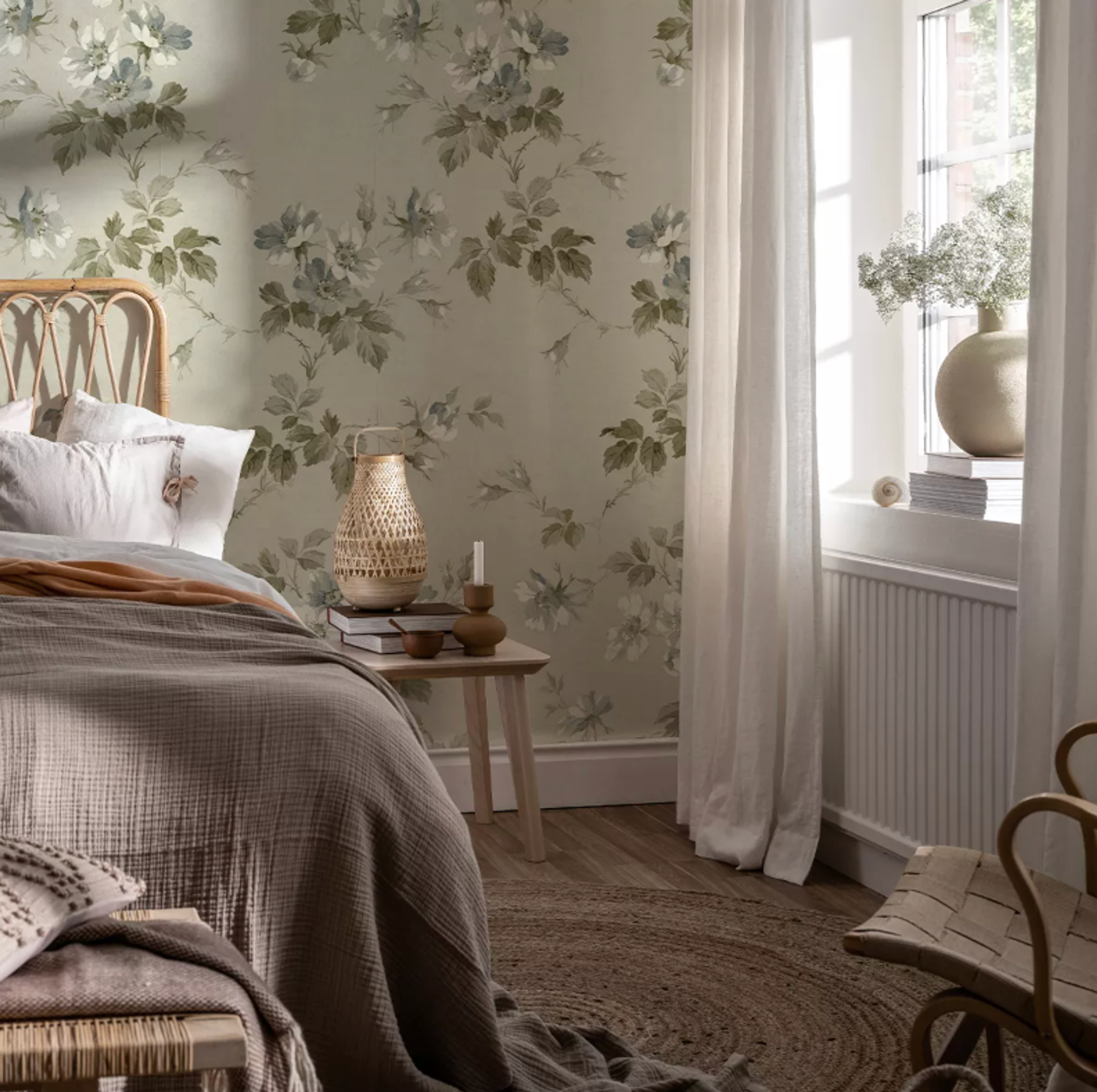 A bedroom with light green floral wallpaper, a rattan bed, a wooden nightstand, and a sunlit window with sheer white curtains.