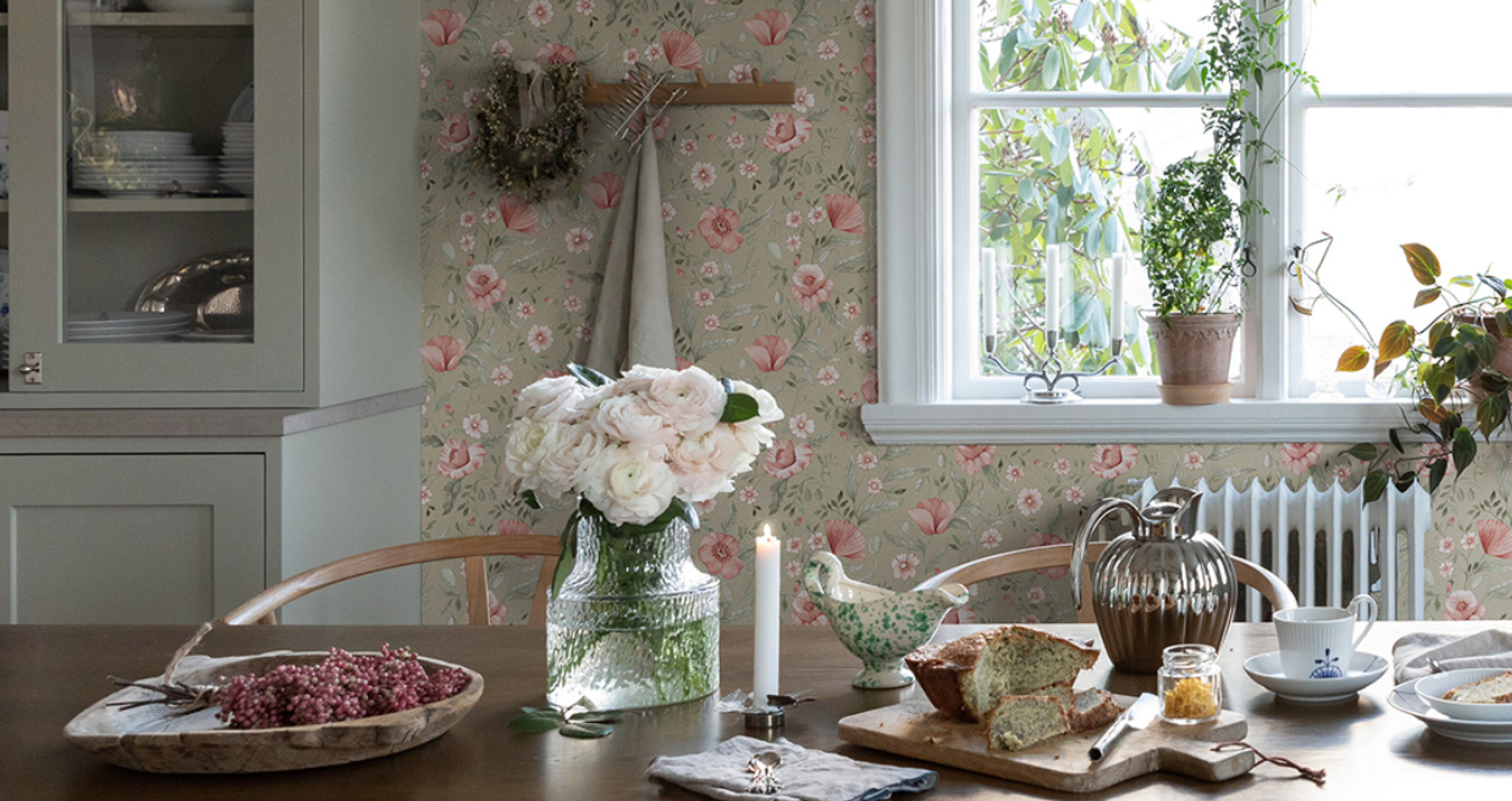 A dining table set with white flowers, bread, and a lit candle, against floral wallpaper and a window.