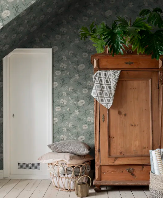 Room interior featuring green floral wallpaper, a wooden wardrobe with a large plant, a white door, and a basket of pillows.