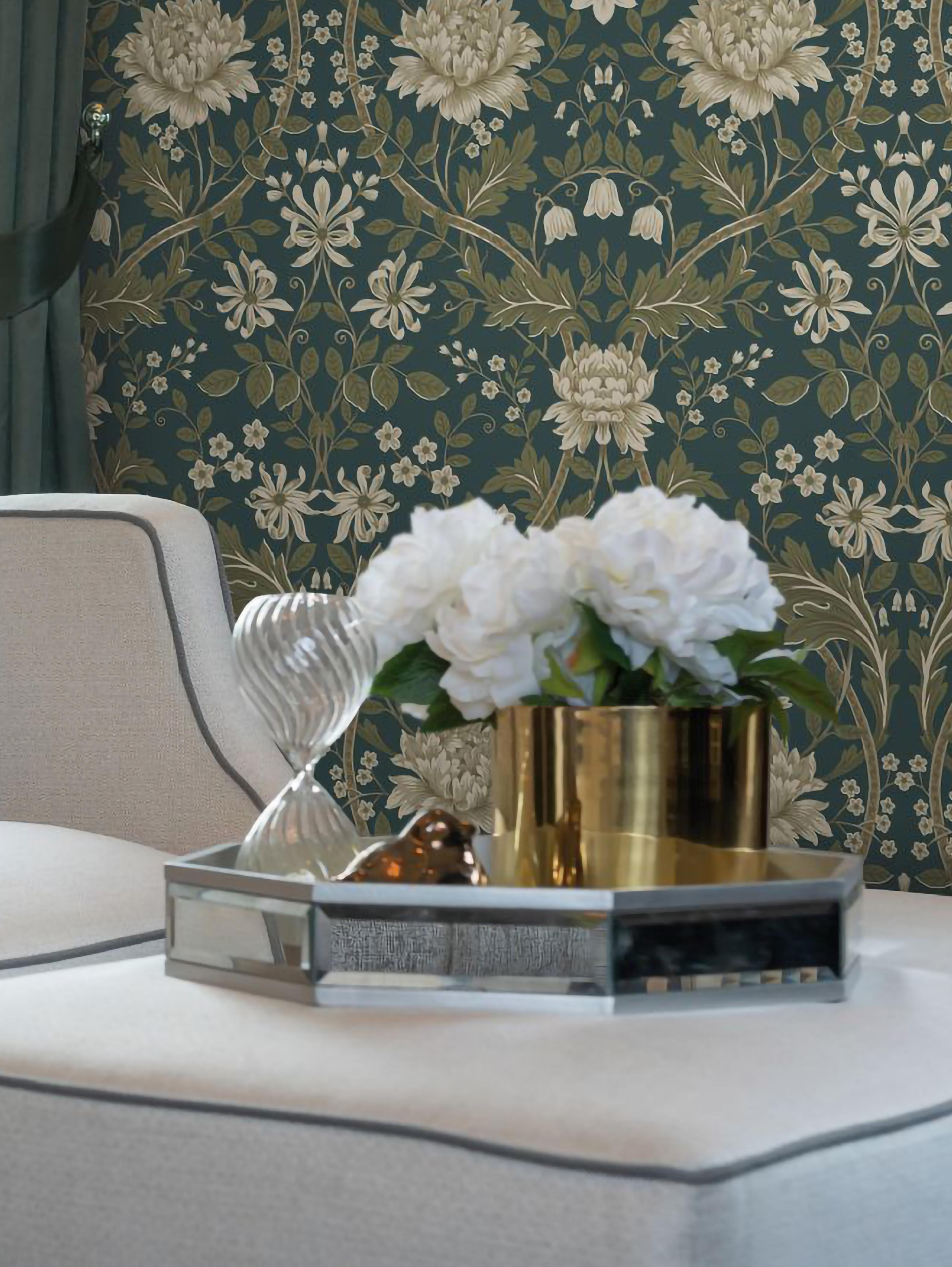Elegant room with dark teal floral wallpaper, a mirrored tray holding white flowers in a golden vase, and a clear glass sculpture on light upholstered furniture.
