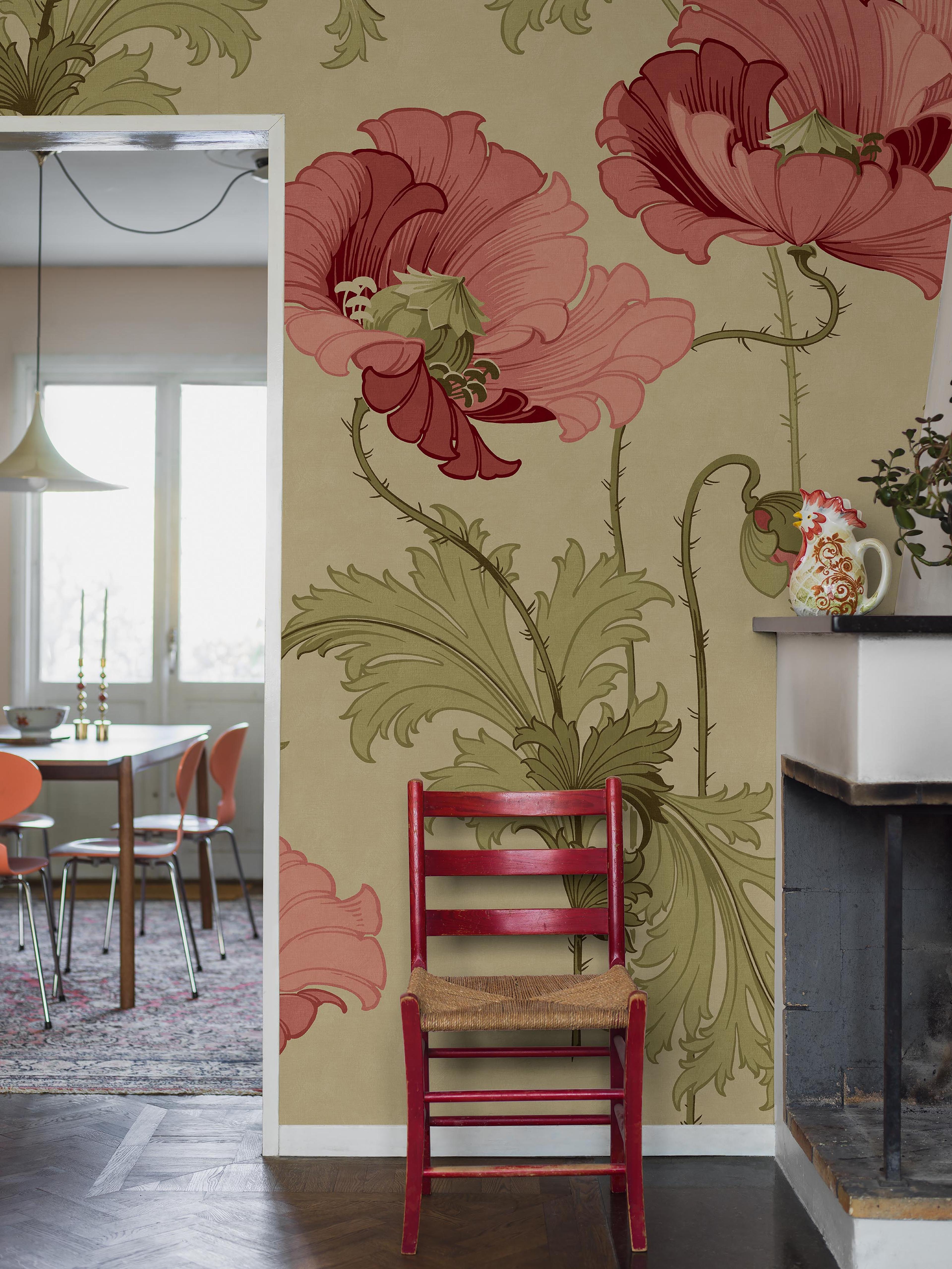 A room with tan wallpaper featuring large pink poppy flowers and green leaves, a red chair with a woven seat, and a dining room visible through a doorway.