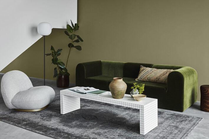 A modern living room with olive green and white walls, featuring a green velvet sofa, a grey sculptural armchair, and a grid-patterned coffee table on a grey rug.