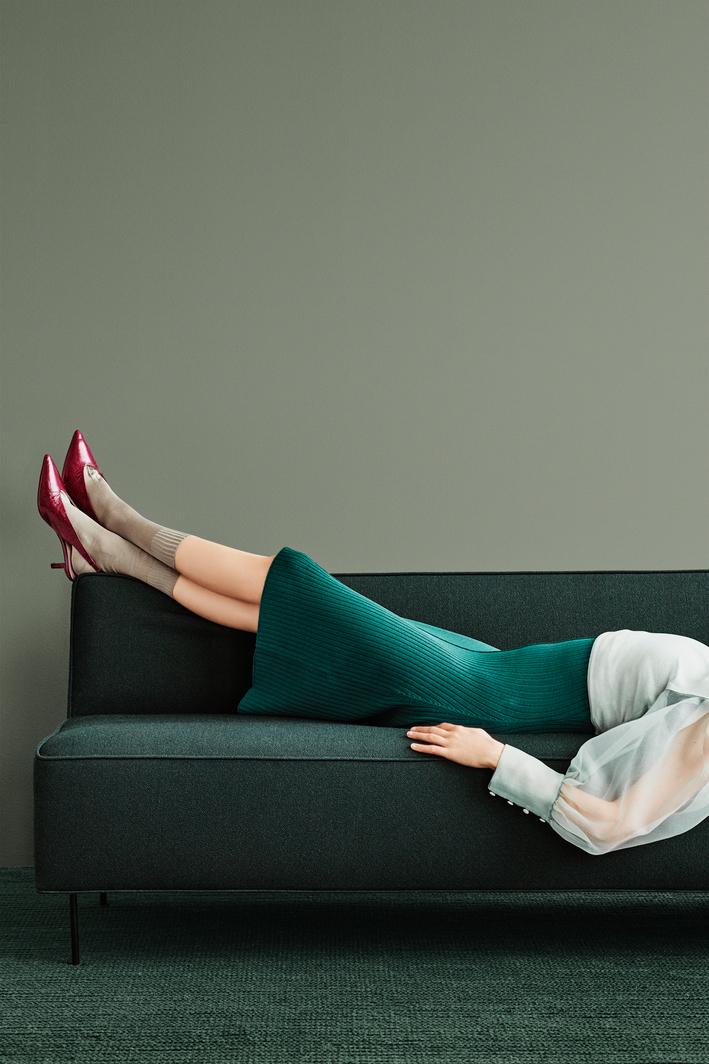A person wearing a green ribbed dress, tan socks, and red high heels lies on a dark green sofa.