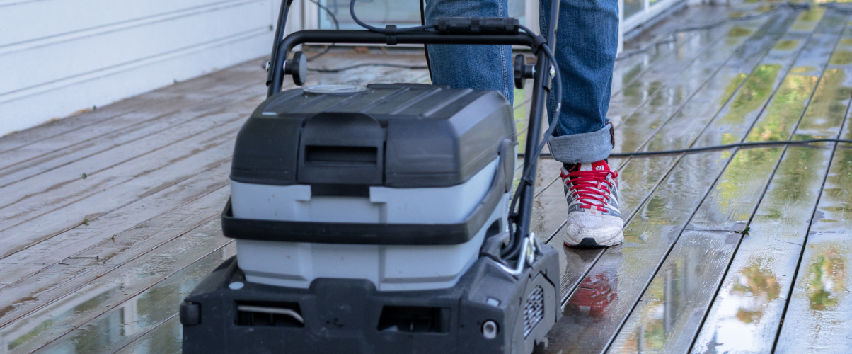 A person uses a surface cleaner to wash a wet wooden deck.