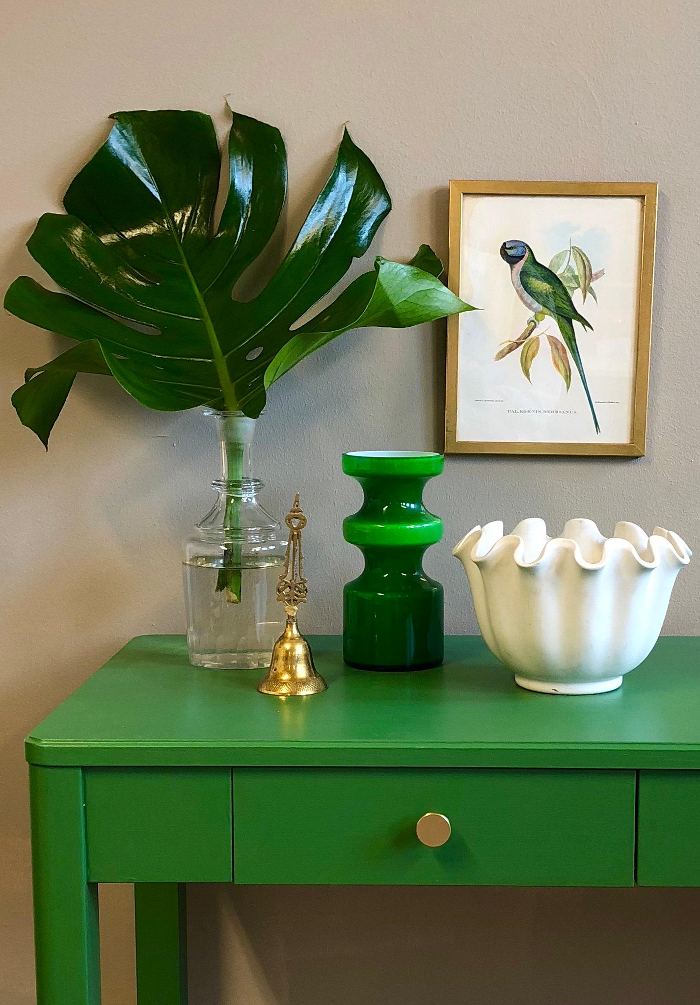 A vibrant green table displays a large monstera leaf in a vase, a brass bell, a green vase, a white ruffled bowl, and a framed parrot print hangs above.