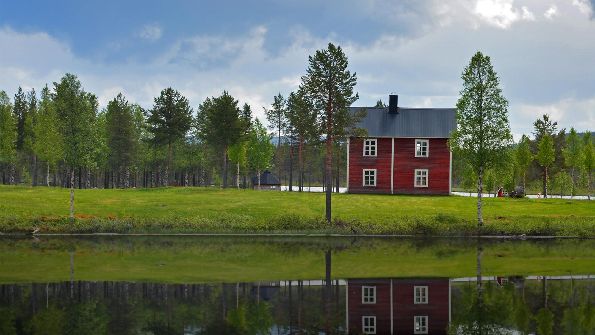A red house on a green hill, backed by a forest, reflected in still water under a cloudy sky.