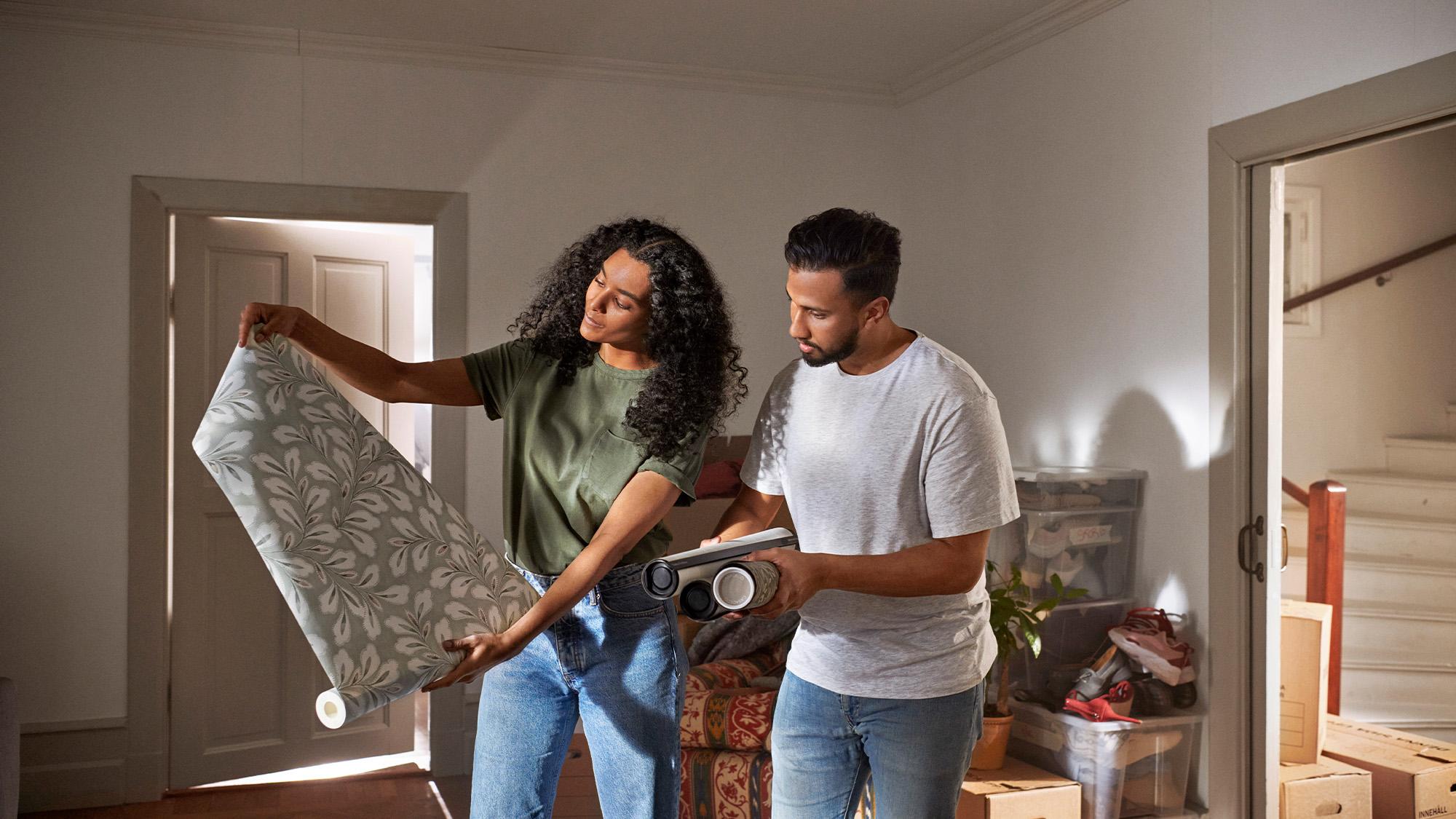 A couple examines wallpaper samples in a room with moving boxes.
