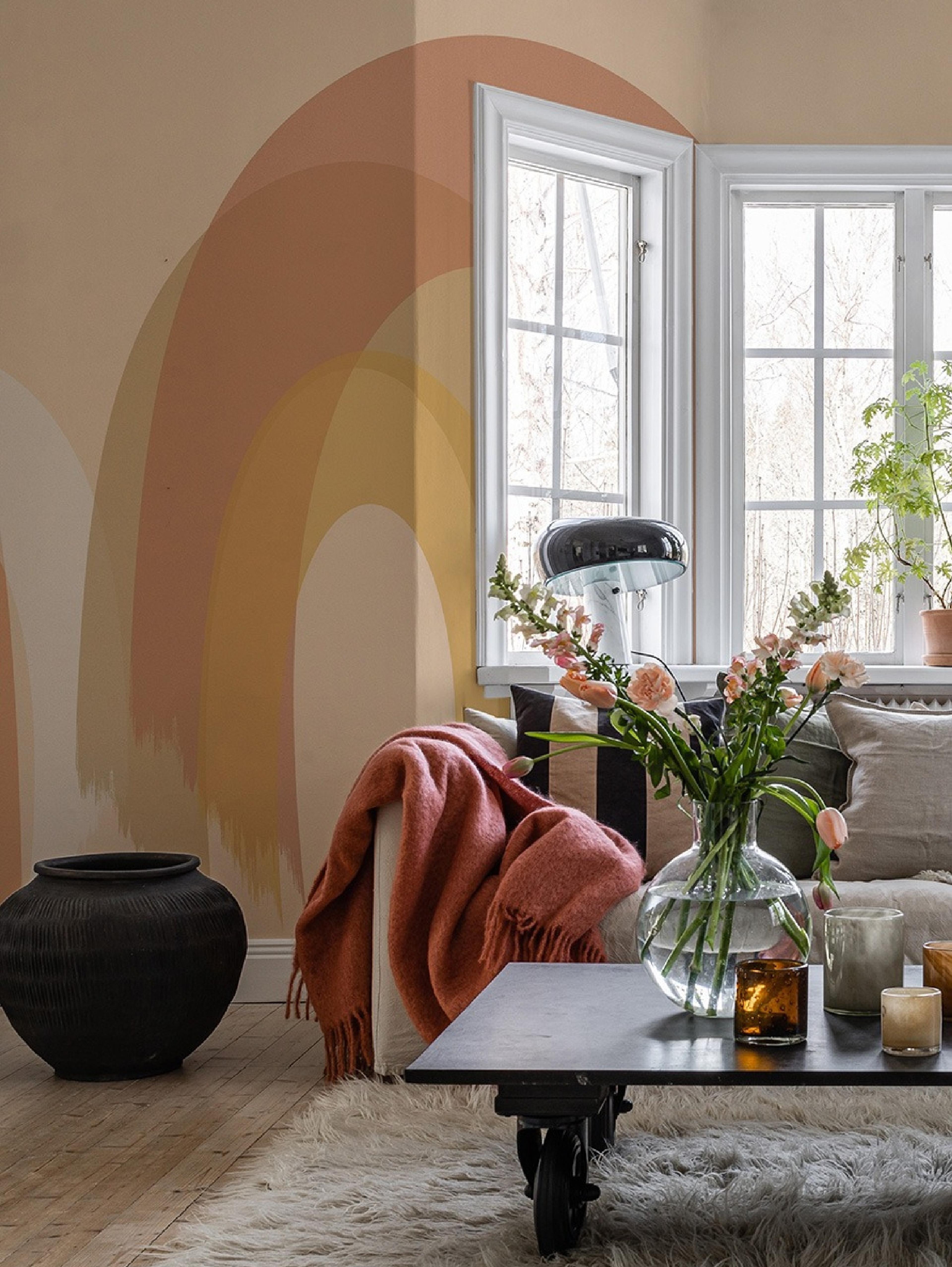 A living room with an earthy-toned abstract rainbow wall mural, a sofa draped with a peach blanket, a coffee table with flowers, and large windows.