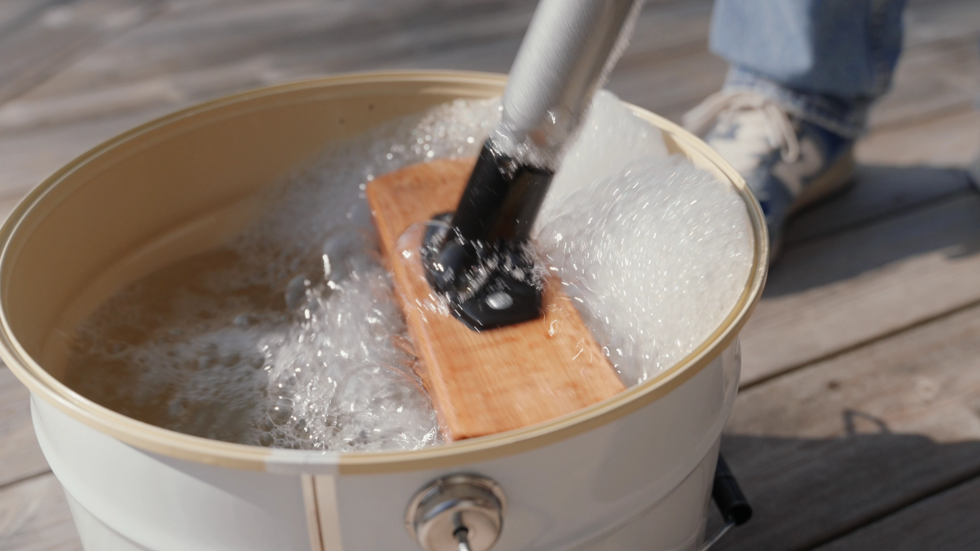 A wooden board is being agitated in a bucket of foamy water on a wooden deck.