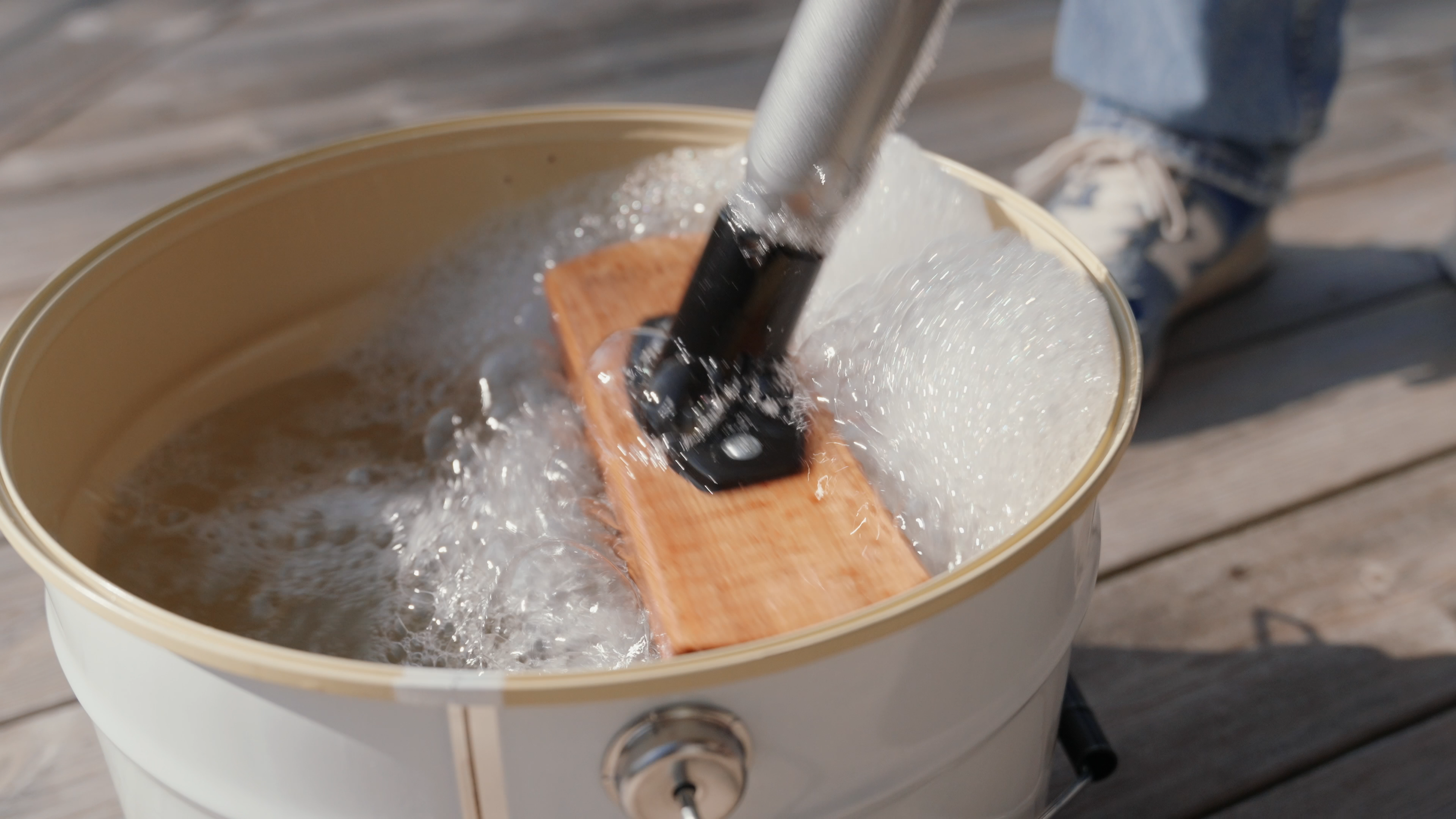 A wooden board is being agitated in a bucket of foamy water on a wooden deck.