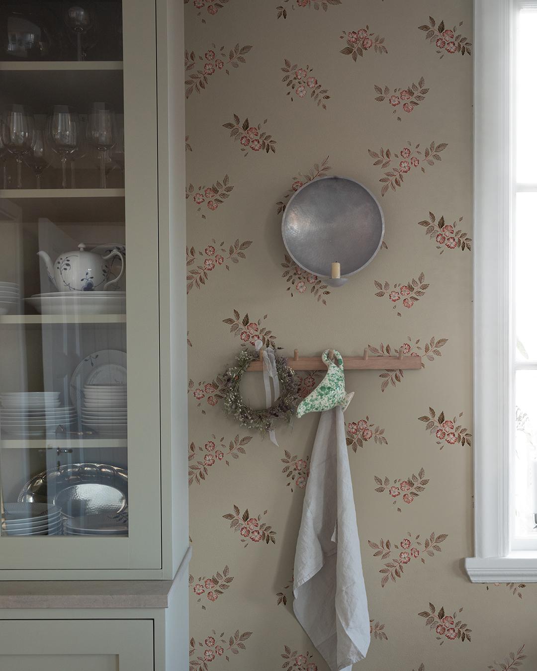 A vintage-style room with a glass-front cabinet next to a wall featuring rose-patterned wallpaper, a metal sconce, and a peg rack with a wreath and towel.