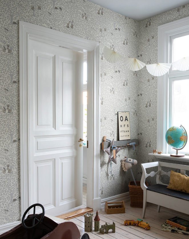 A light-filled child's room with forest animal wallpaper, a white door slightly ajar, a bench, a globe, and various toys on a pale wooden floor.