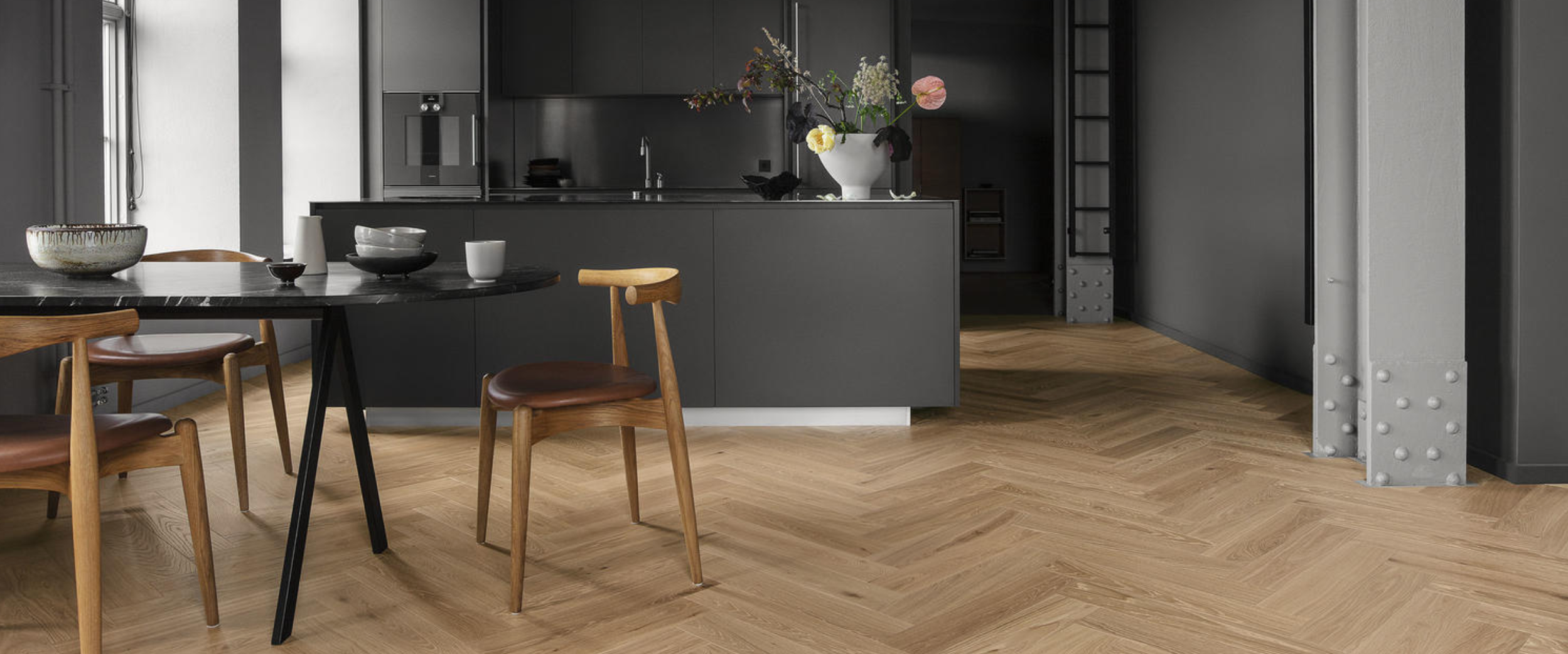 Modern open-plan kitchen and dining area with dark gray cabinetry, a black table, wooden chairs, and light herringbone wood floors.