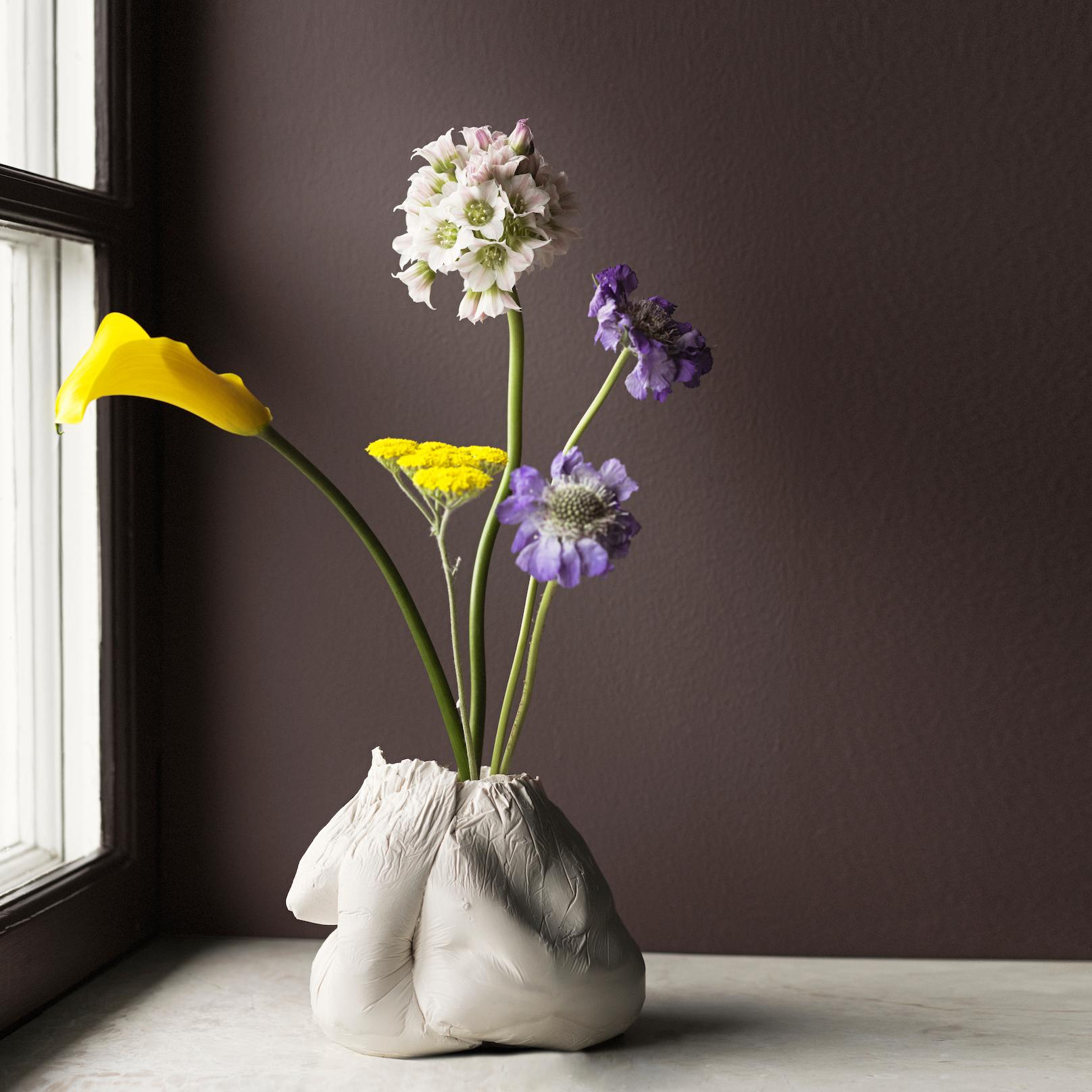 A textured white vase holds yellow, white, and purple flowers on a counter by a window.