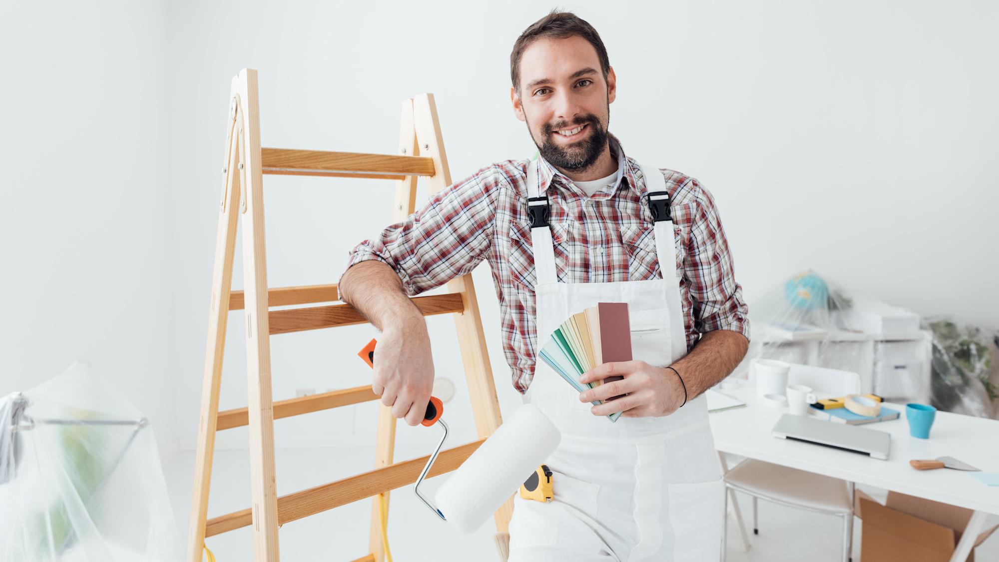 Smiling painter in overalls holding a roller and color swatches, leaning on a ladder.