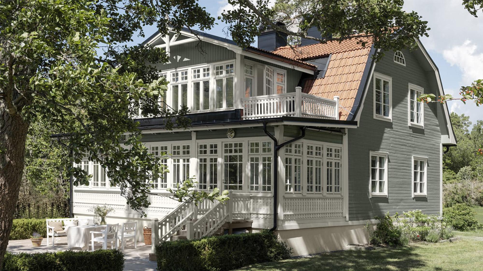 A light green house with white trim features a sunroom, an upper balcony, and a patio with white outdoor furniture, surrounded by trees.