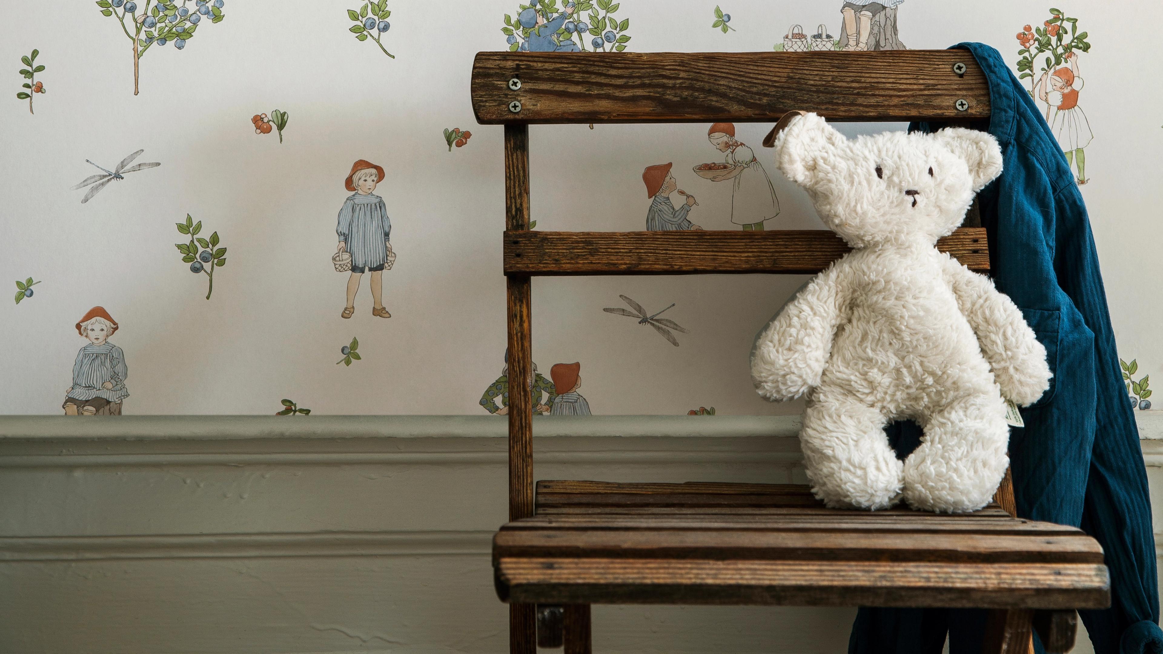 A fluffy white teddy bear sits on a rustic wooden chair with a blue blanket, in front of whimsical wallpaper depicting children and blueberry bushes.
